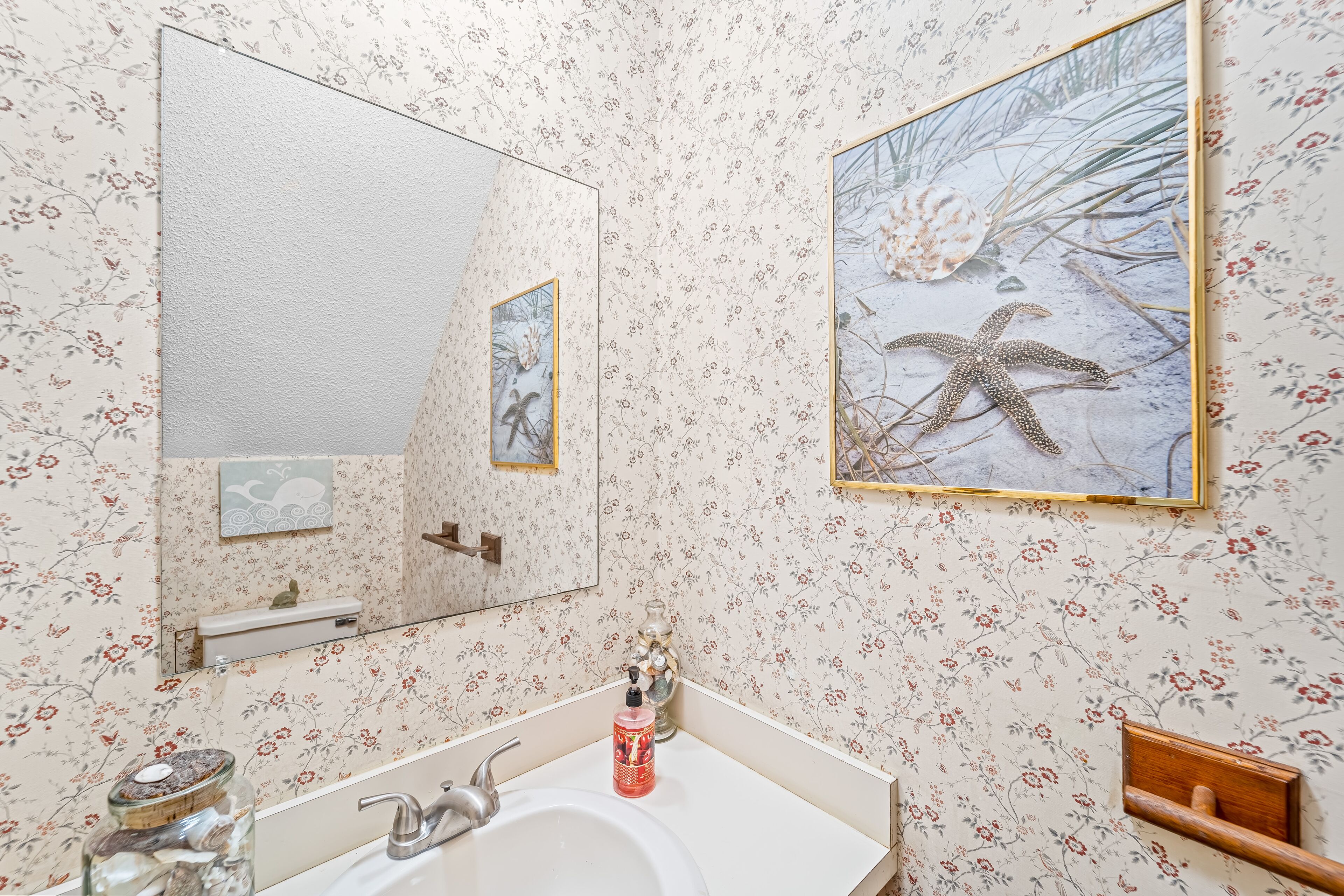 A bathroom with floral wallpaper, a sink with a mirror, and two framed pictures on the wall.