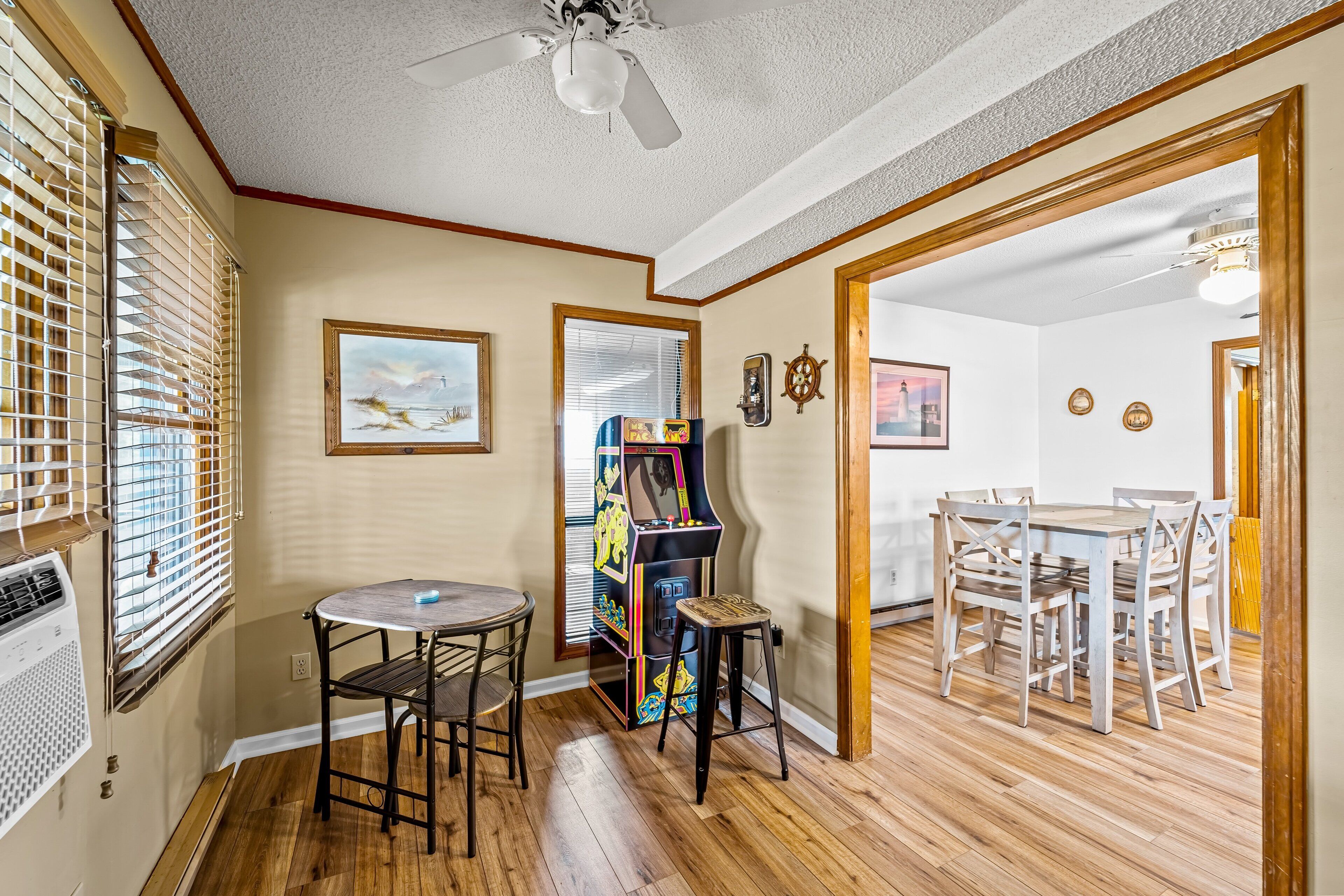A room with a wooden floor, a ceiling fan, a table with chairs, a small stool, and a colorful arcade machine.