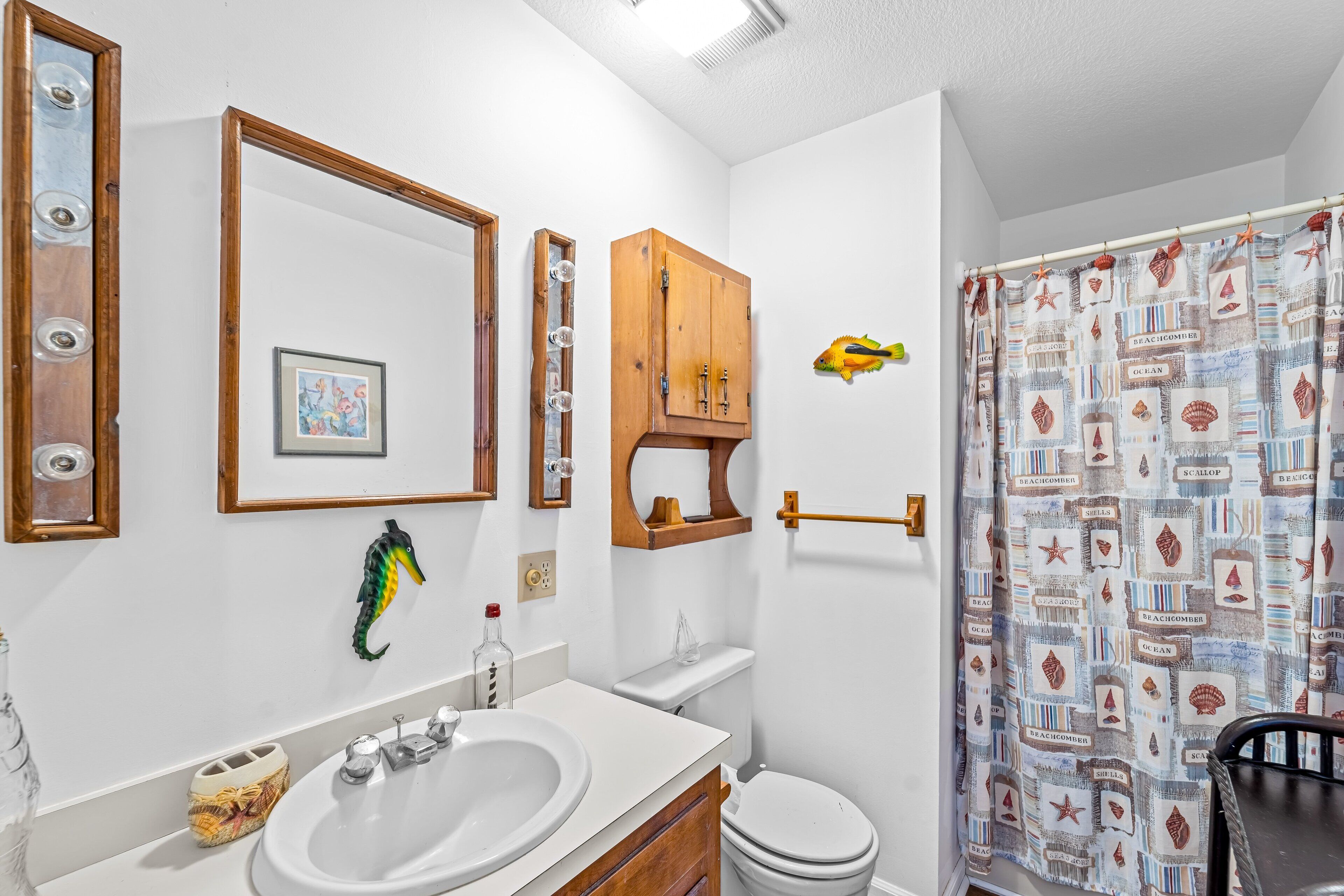 Un bagno con lavandino bianco, uno specchio, un mobiletto in legno, un WC e una tenda da doccia a tema spiaggia.