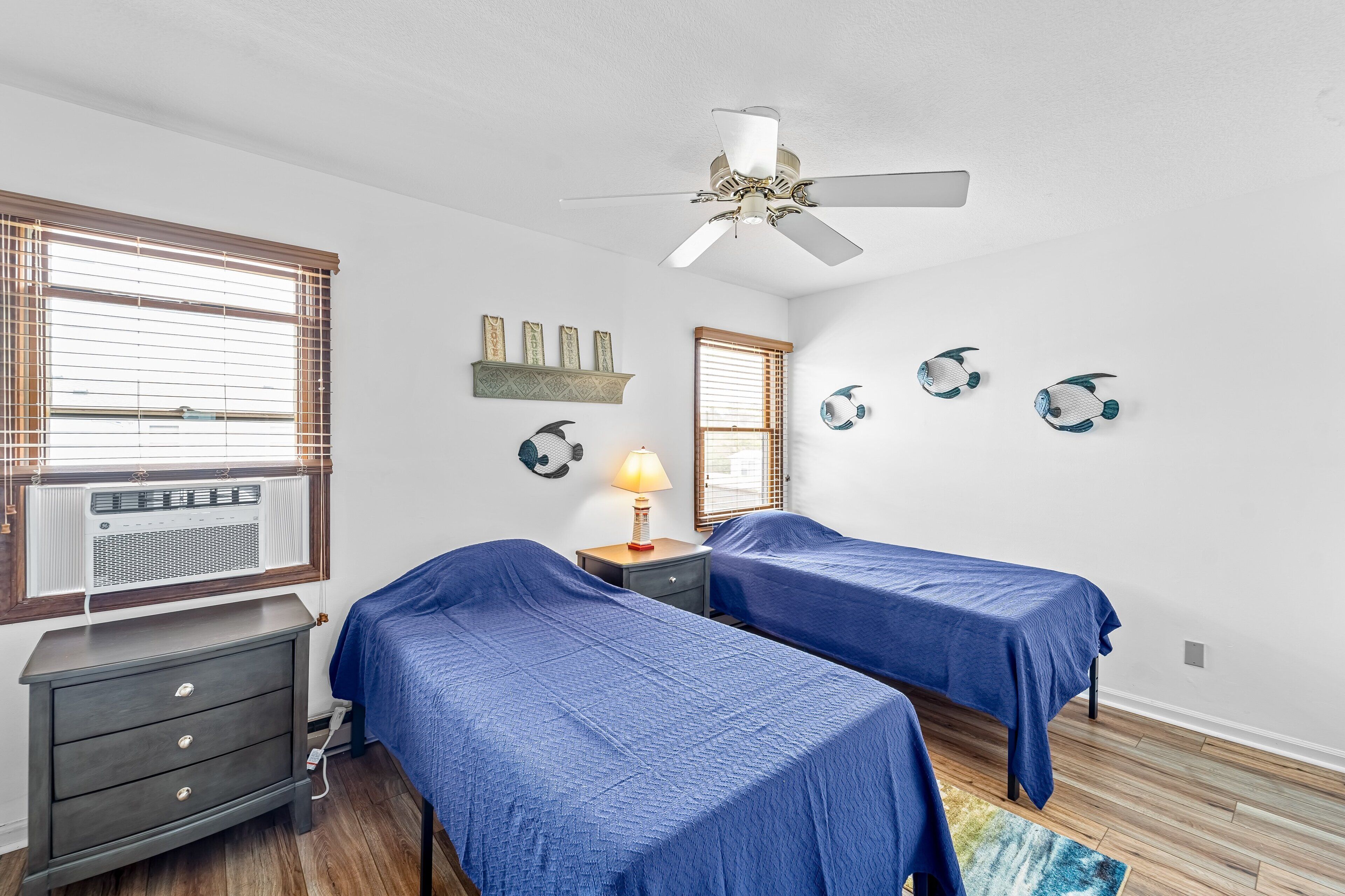 A bedroom with two beds, a ceiling fan, a nightstand with a lamp, and wall decorations.