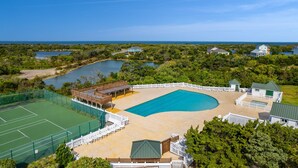 A large outdoor swimming pool surrounded by a wooden deck, tennis courts, and a white fence.
