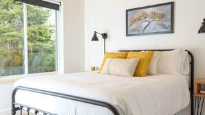 A bed with white bedding and yellow pillows, a black metal headboard, a bedside table with a lamp, and a painting of a tree on the wall. - Forest Cabin w/ Stunning Wall of Windows (Jackson)