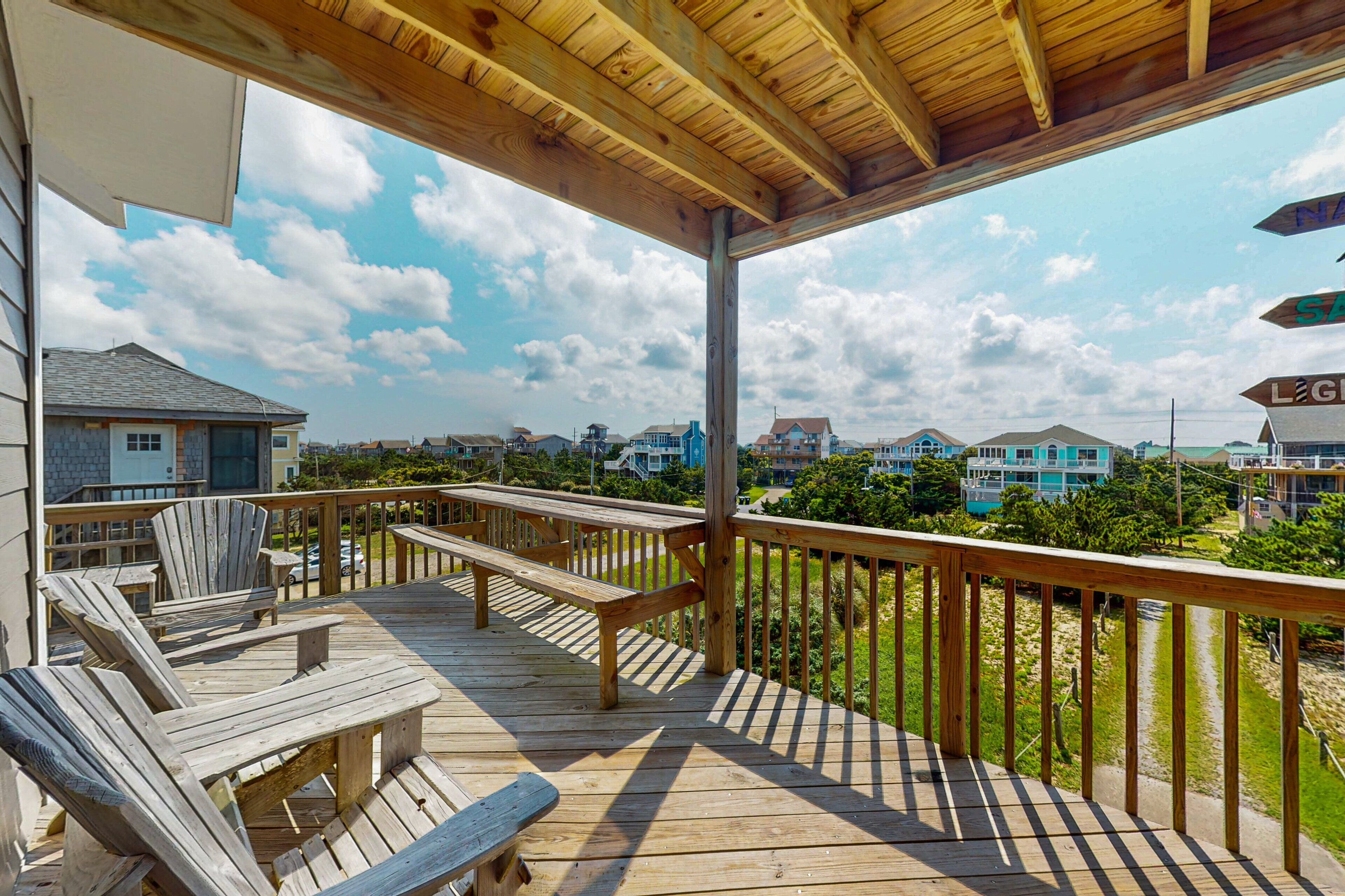 A wooden deck with Adirondack chairs and picnic tables, overlooking a residential area with houses and a clear sky.