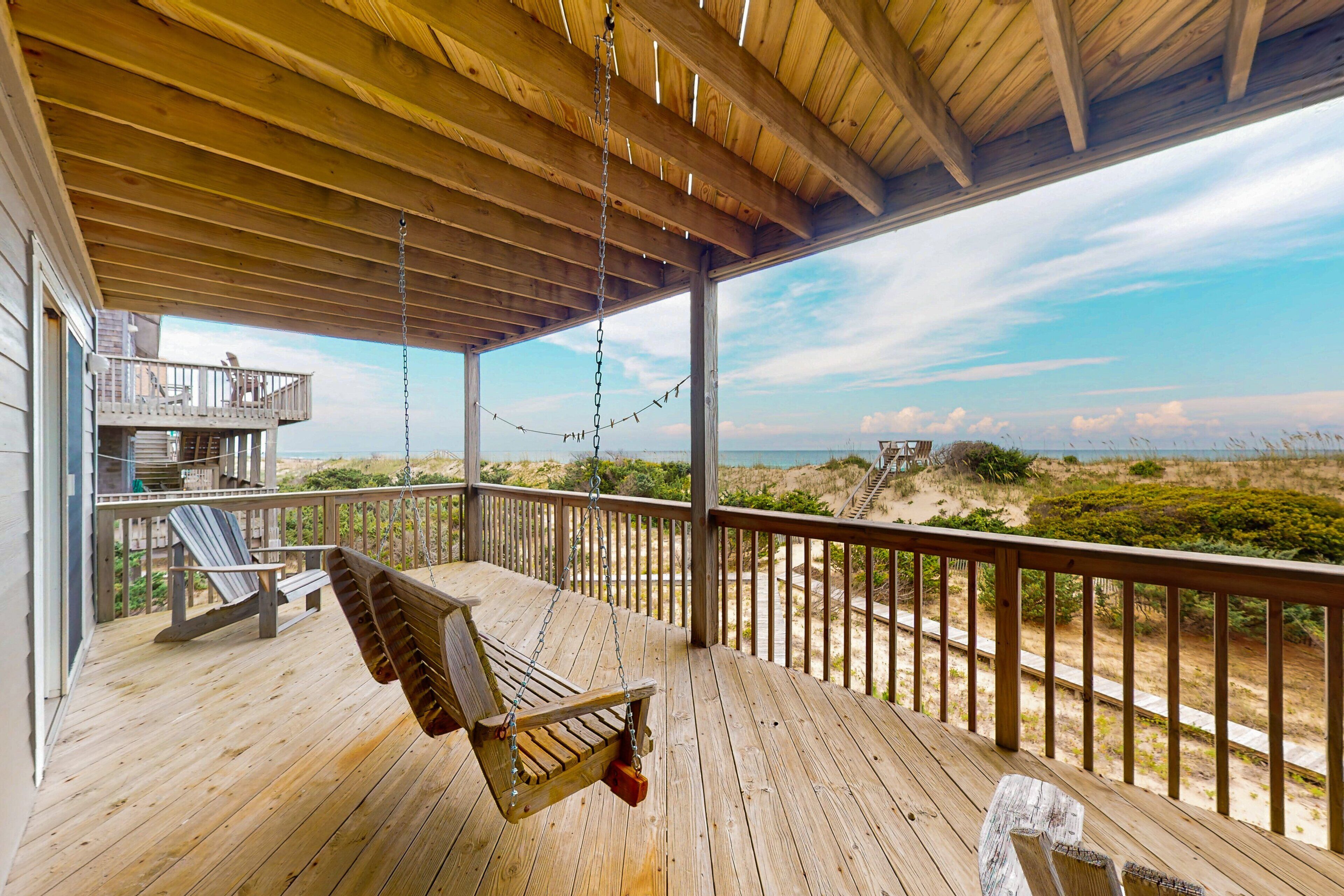 A wooden deck with a swing chair, a bench, and a view of the beach.