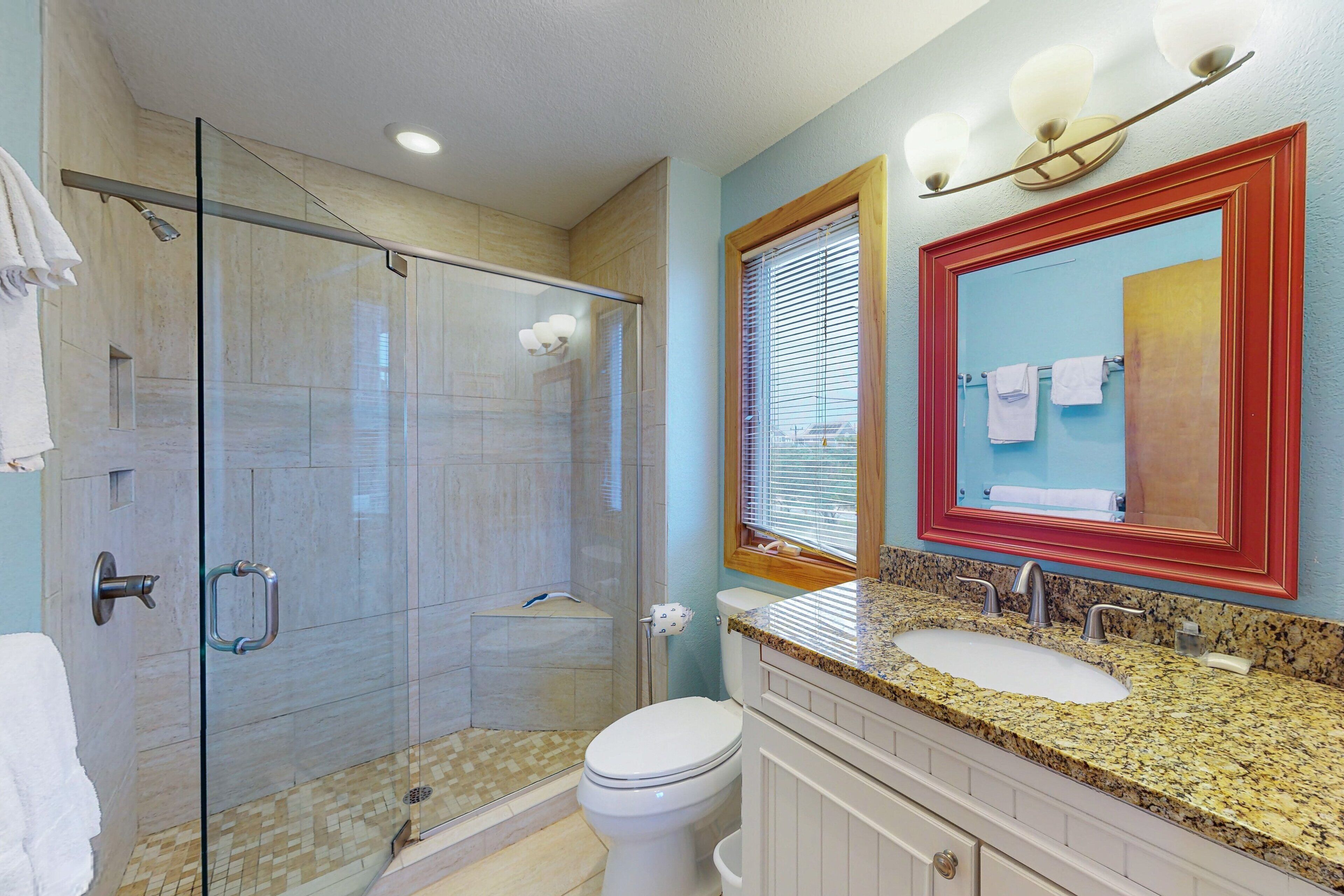 A bathroom with a glass-enclosed shower, a white toilet, a sink with a granite countertop, and a mirror with a red frame.