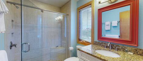 A bathroom with a glass-enclosed shower, a white toilet, a sink with a granite countertop, and a mirror with a red frame.