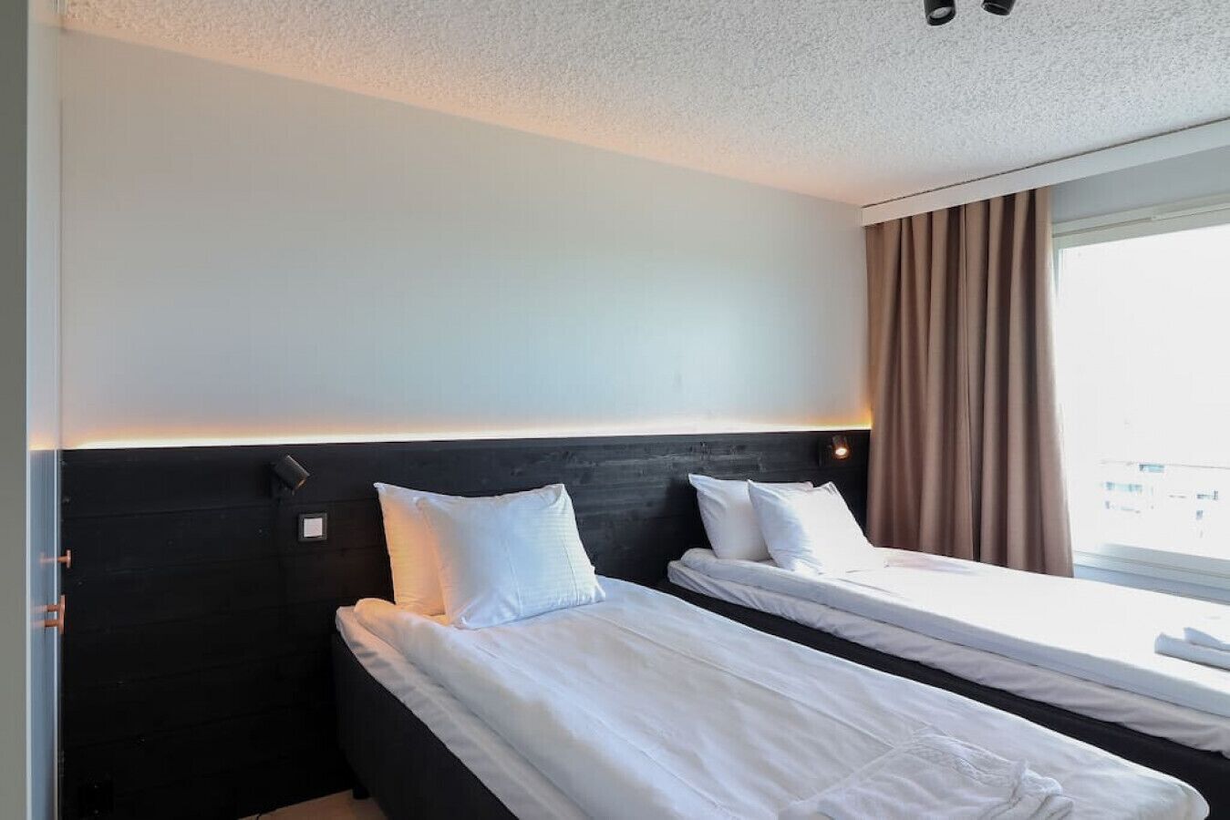 A hotel room with two single beds, a dark headboard, a window with curtains, and a view of the city.