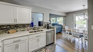 A modern kitchen with white cabinets, a dishwasher, and a coffee maker. There is a dining area with white chairs and a blue couch. - Charming 3BR home in Frisco with Wifi, Full Kitchen and Private Hot Tub (Frisco)