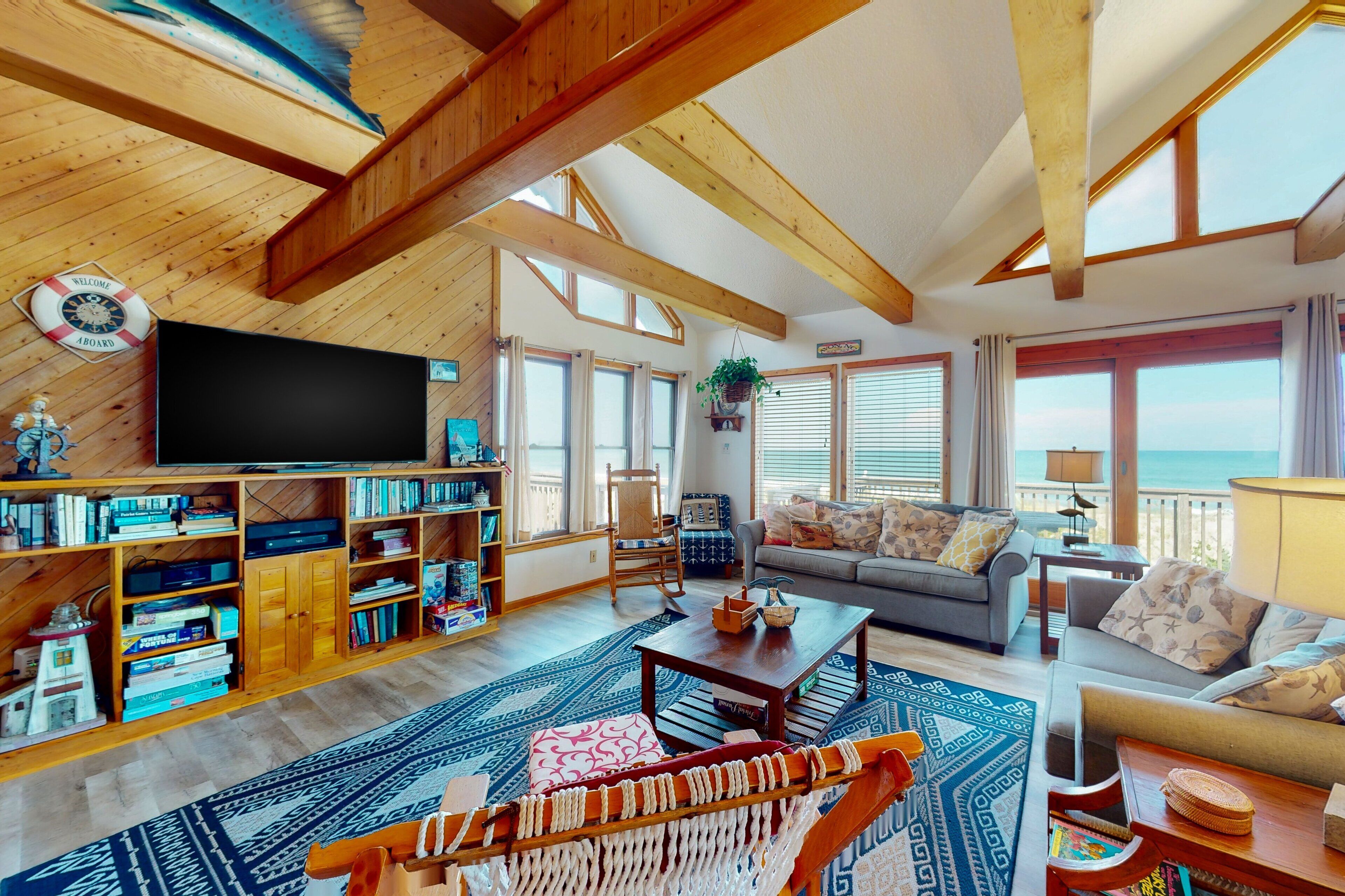 A spacious living room with a large TV, wooden bookshelves, a sofa, and a view of the ocean.