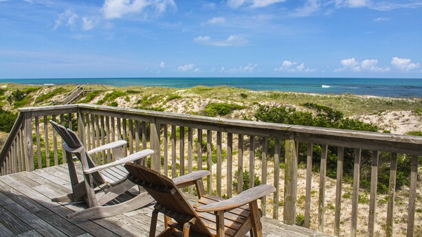 Eine Holzterrasse mit zwei Adirondack-Stühlen mit Blick auf einen Strand, Dünen und das Meer.