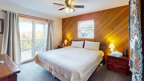 Een slaapkamer met een groot bed, houten wandpanelen, een plafondventilator, een nachtkastje met een lamp en uitzicht door een schuifdeur.