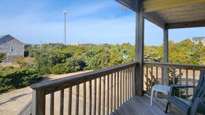 Une terrasse en bois avec une balustrade, une chaise en plastique blanche et une vue sur une zone boisée, avec une grande structure visible au loin.