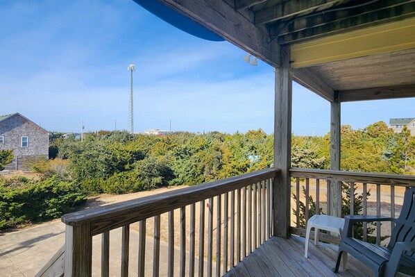 A wooden deck with a railing, a white plastic chair, and a view of a forested area with a tall structure in the distance.