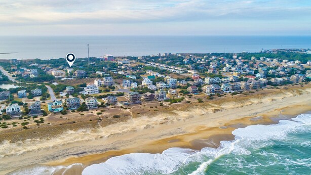 Vue aérienne d’une zone résidentielle côtière, avec des maisons le long de la plage, un ciel dégagé et l’océan au premier plan.