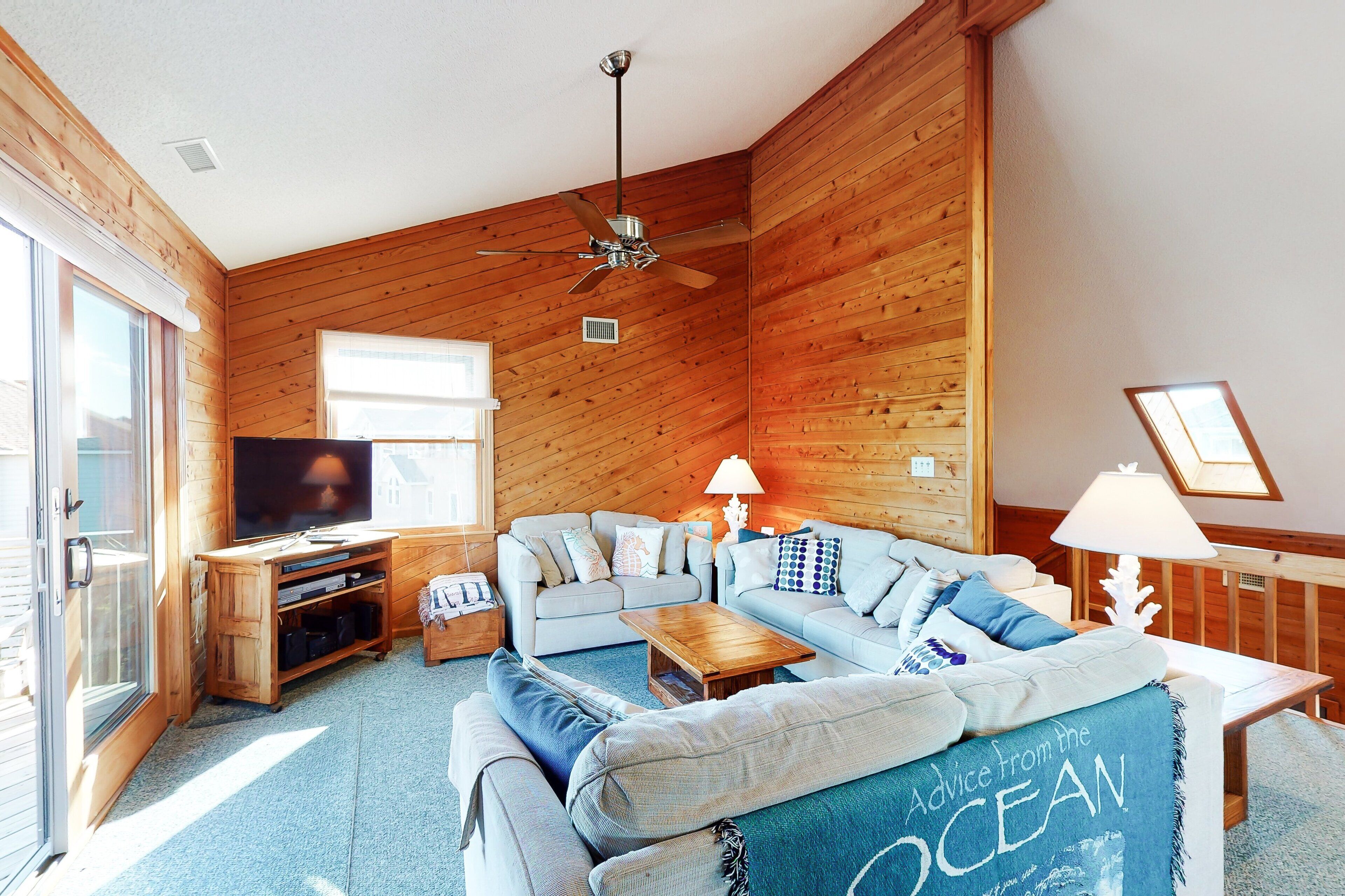 A cozy living room with a wooden interior, a ceiling fan, a flat-screen TV, and a large window.