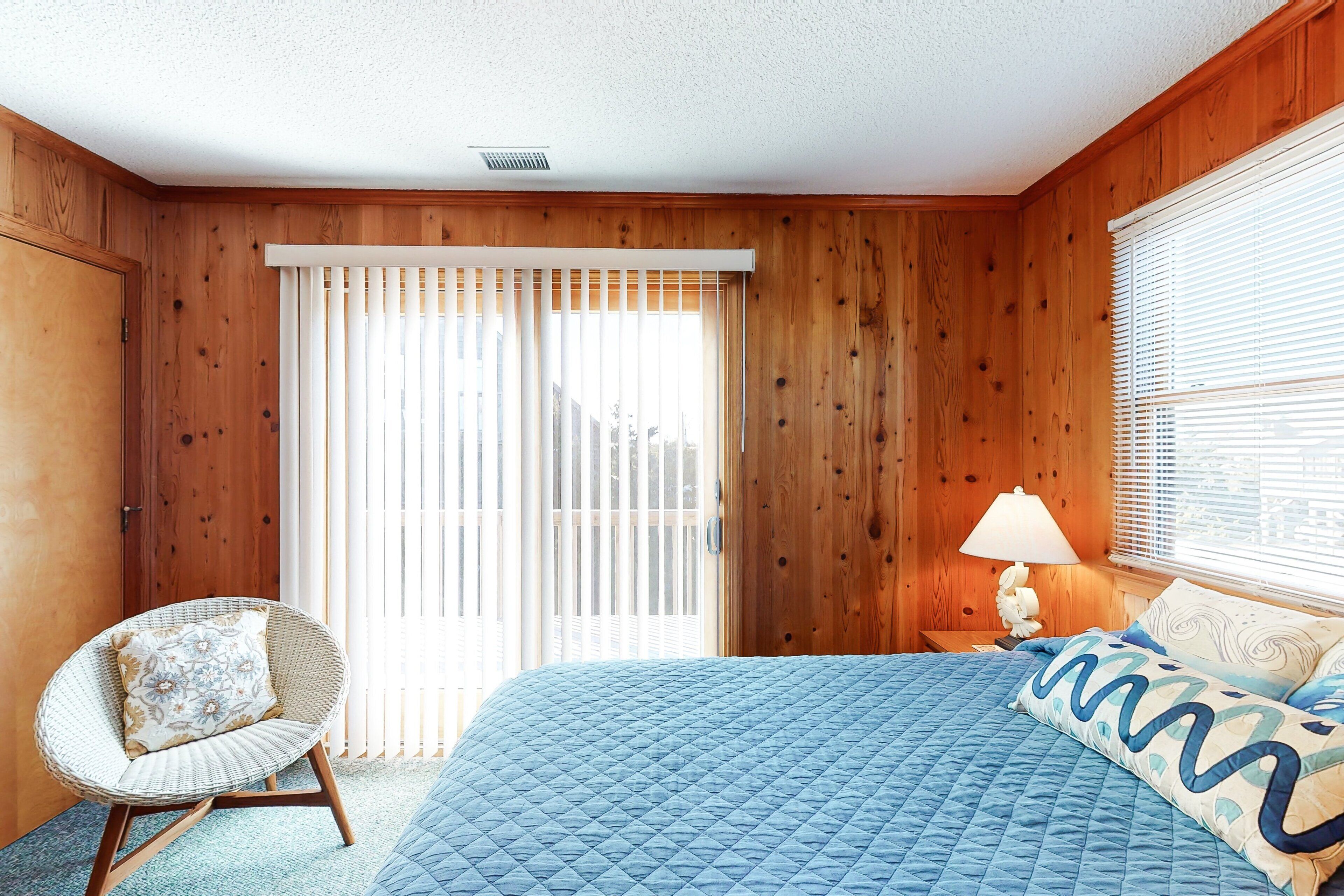 Une chambre avec des murs en bois, un lit avec une literie bleue, une chaise en osier agrémentée d’un coussin à motifs floraux et une fenêtre avec des stores.
