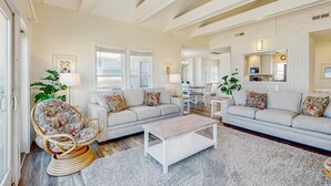 A spacious living room with a light-colored sofa, a wicker chair, and a coffee table. There are two windows providing natural light, and a kitchen area is visible in the background. - Fun beachfront home w/shared pool, WiFi, private washer & dryer, central AC (Avon)