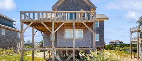 A wooden house with a balcony on stilts, surrounded by beach vegetation.