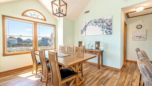 A dining area with a wooden table and chairs, large windows, and artwork on the walls. - Semi-Ocean Front Retreat with Full Kitchen, Wifi, Outdoor Shower and A/C (Avon)