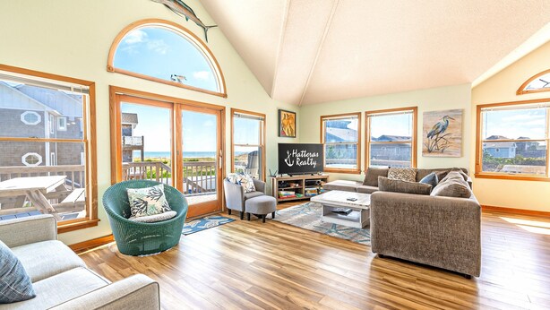 A spacious living room with a large window overlooking a scenic view, a cozy seating area, and a 'Hatteras Realty' sign on the wall. - Semi-Ocean Front Retreat with Full Kitchen, Wifi, Outdoor Shower and A/C (Avon)