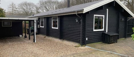 A black wooden building with a sloped roof, a covered porch, and a gravel driveway.