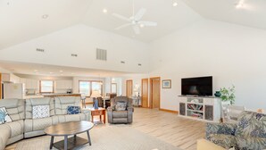 A spacious living area with a sofa, armchair, and coffee table. A television is mounted on the wall, and there is a kitchen in the background. - Oceanfront home with central AC, ocean view, private hot tub, & pool (Avon)