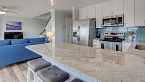 A modern kitchen with white cabinets, a stainless steel refrigerator, a microwave, and a stove. There is a blue sofa and a dining area with a marble countertop. - Stunning beachfront home with ocean-view decks and direct beach access in Surf City (Surf City)