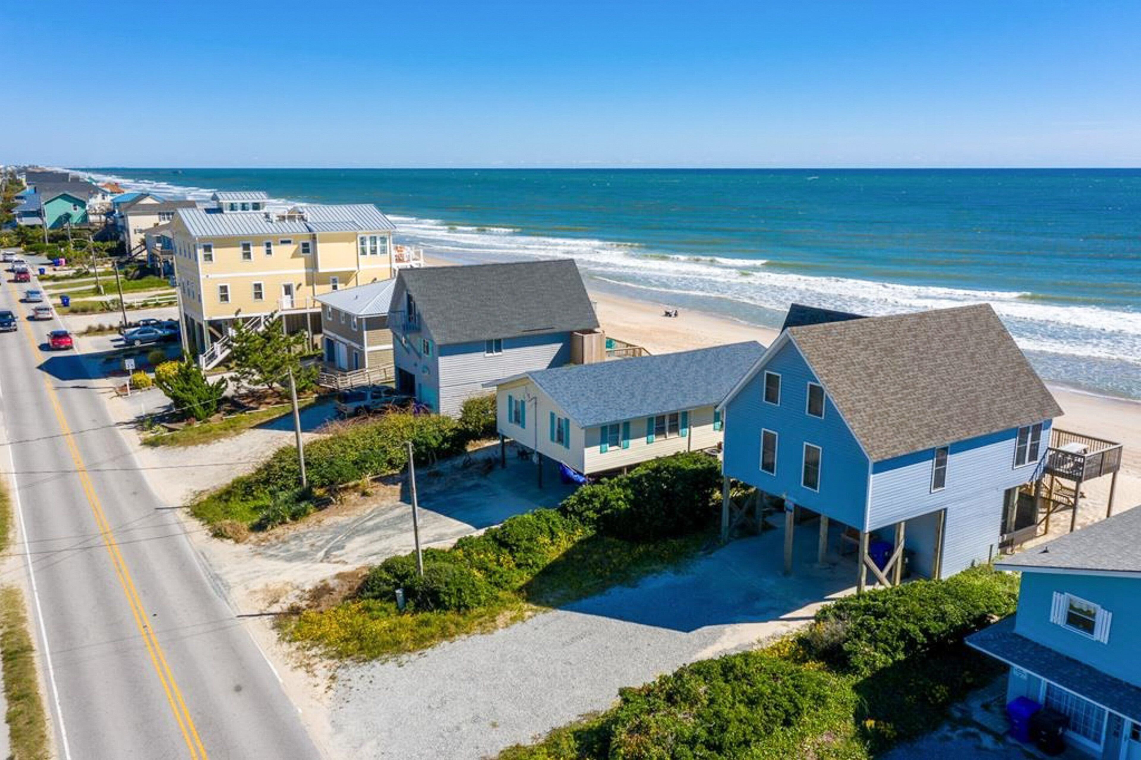 A coastal residential area with houses near the beach, a road, and a clear view of the ocean.