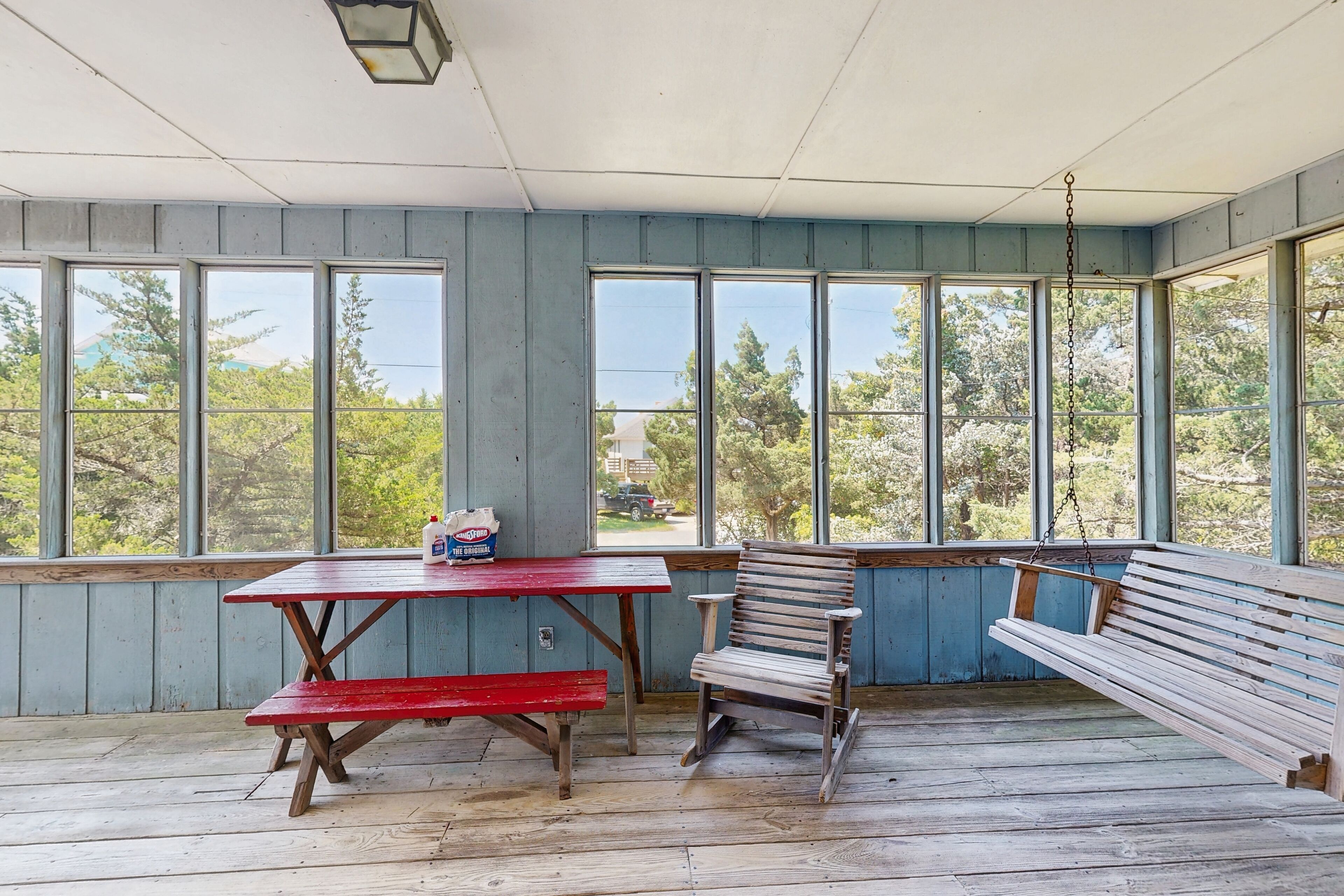 Une terrasse couverte avec un banc en bois, une table de pique-nique et de grandes fenêtres donnant sur les arbres et les bâtiments.