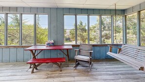 Une terrasse couverte avec un banc en bois, une table de pique-nique et de grandes fenêtres donnant sur les arbres et les bâtiments.