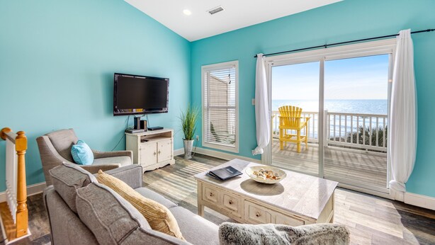 A living room with a television, sofa, coffee table, and a view of the ocean through large sliding doors. - Beachfront home with beach access, community pool, foosball table, ocean views (Surf City)