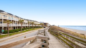 A wooden boardwalk leading to beachfront hotels with balconies and palm trees. - Beachfront home with beach access, community pool, foosball table, ocean views (Surf City)