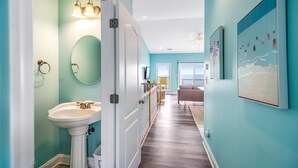 A hallway with a white door leading to a bathroom with a sink and mirror. There is a staircase with a railing and a view of a balcony with a railing. - Beachfront home with beach access, community pool, foosball table, ocean views (Surf City)