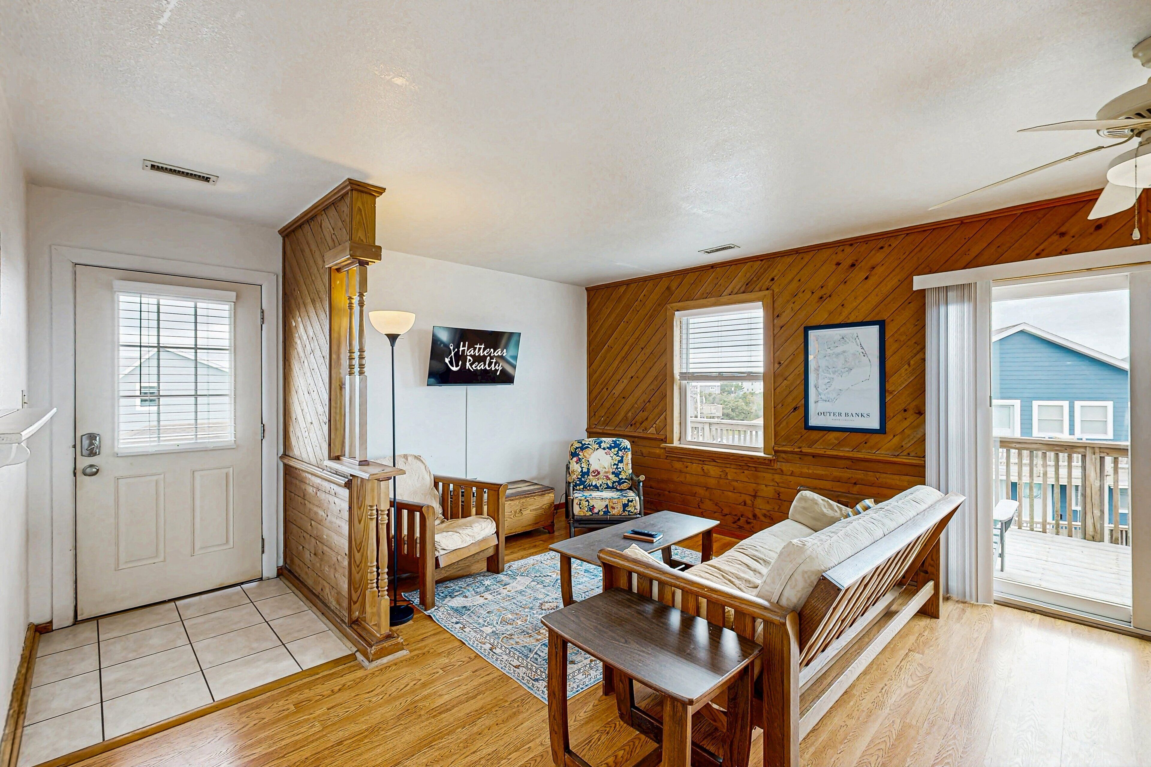 A cozy living room with a wooden bench, a small table, and a chair. There is a door with a window, a wall-mounted sign, and a view of the outside.