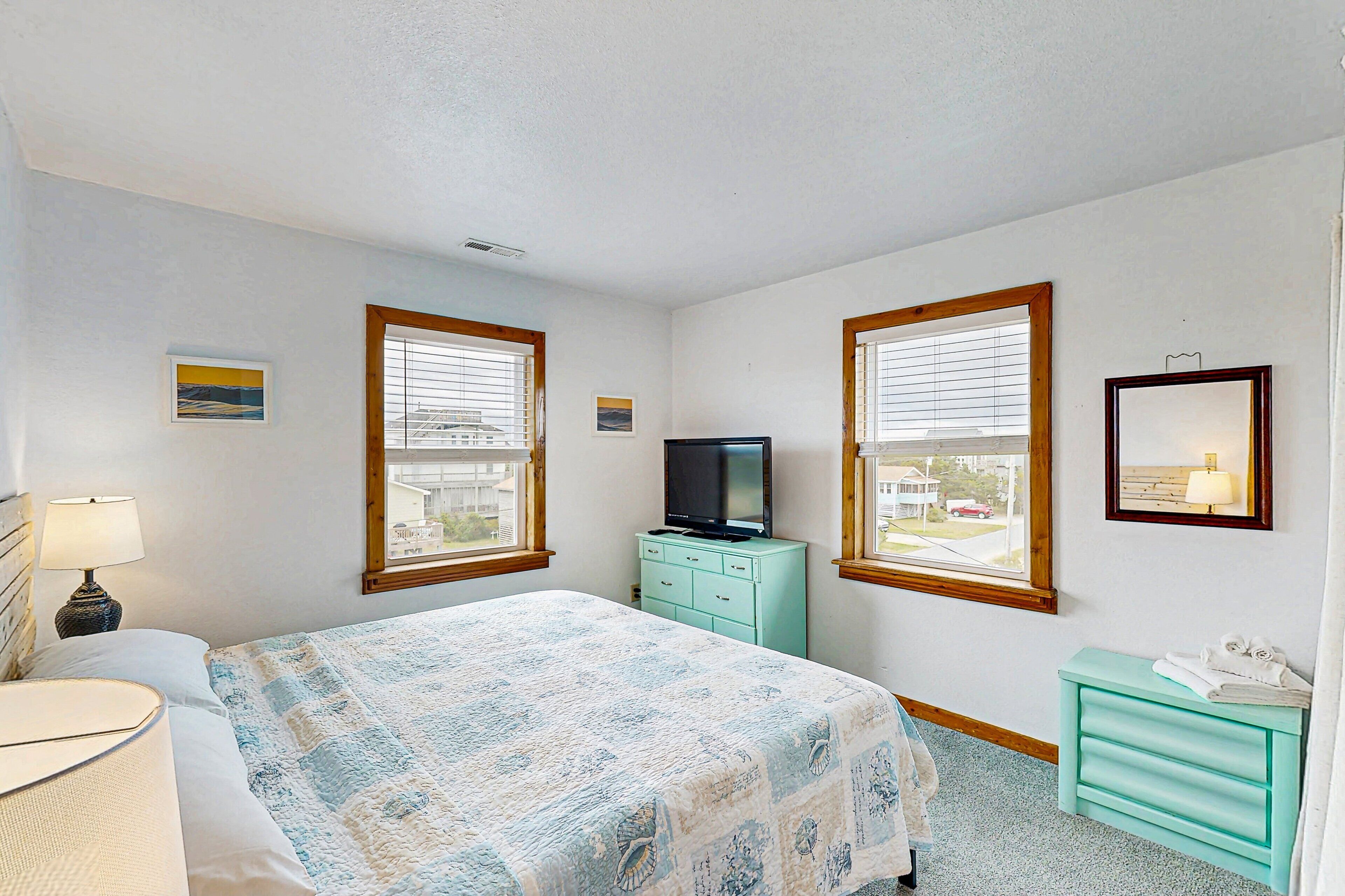 A bedroom with a bed, a dresser, a television, and two windows with blinds.