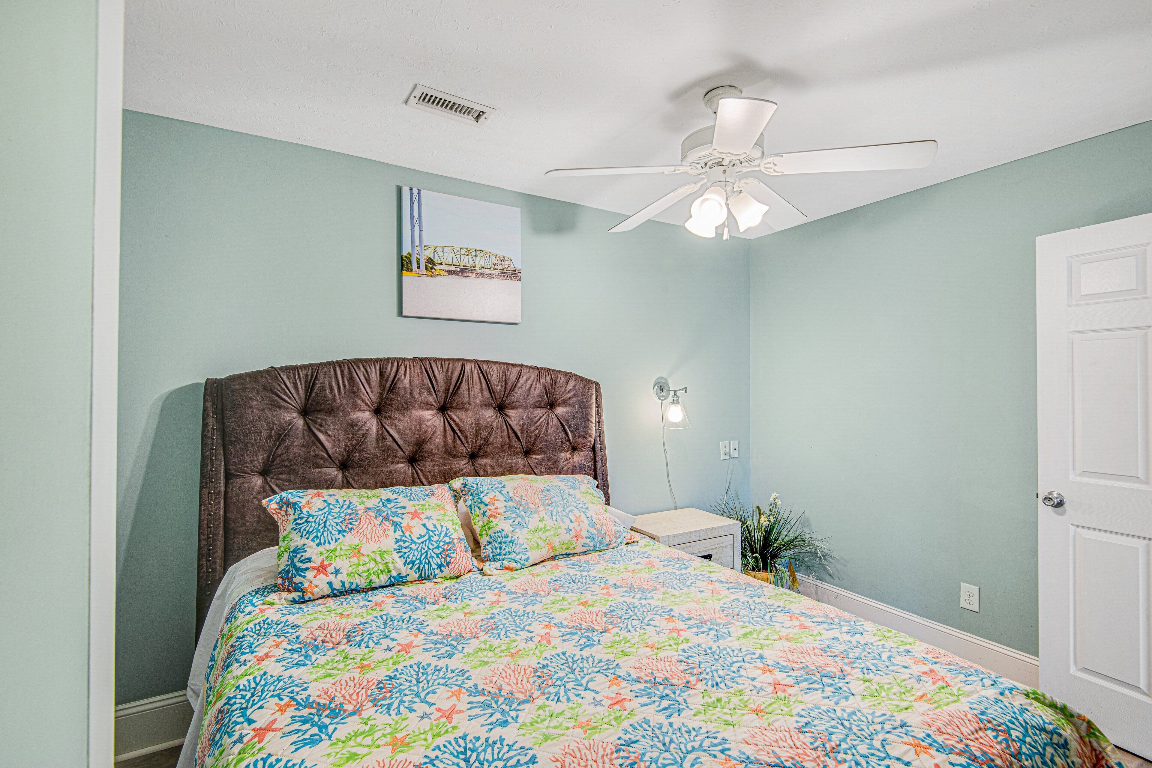 A bedroom with a tufted headboard, a bed with a colorful floral bedspread, a ceiling fan, and a wall-mounted picture.