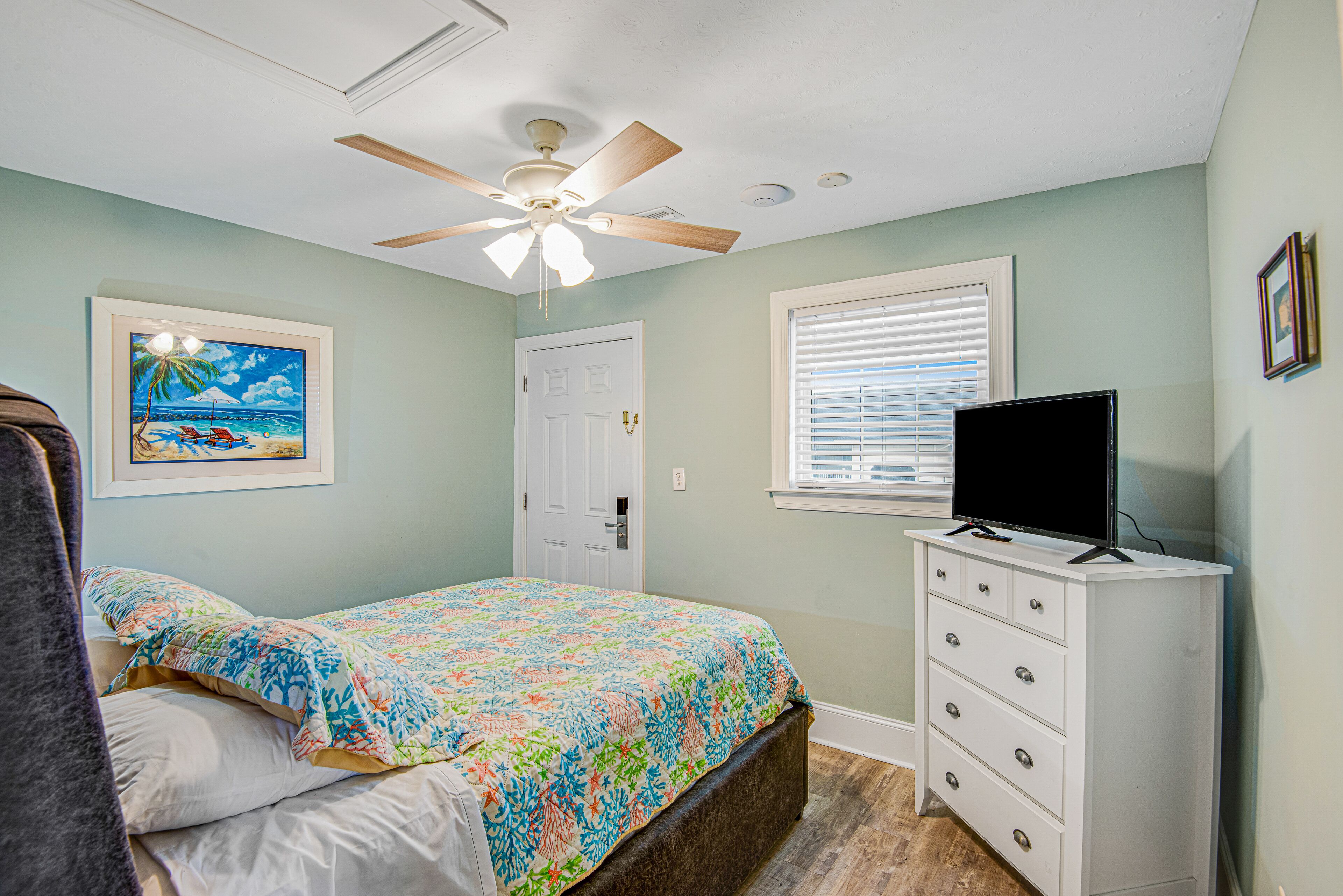 A bedroom with a bed, a ceiling fan, a dresser, a television, a door, and a window with blinds.