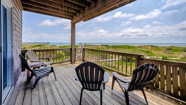 A wooden deck with two Adirondack chairs and a small table, overlooking a beach and dunes. - Bright, updated oceanfront home with shared pool, private deck, & great views (Avon)