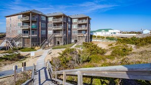 A multi-story building with balconies, a wooden deck, and a sandy area in front. - Bright, updated oceanfront home with shared pool, private deck, & great views (Avon)