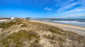 A beach with a wooden pier extending into the ocean, sandy dunes, and a few buildings in the background. - Bright, updated oceanfront home with shared pool, private deck, & great views (Avon)