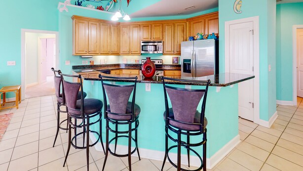 A kitchen with a turquoise color scheme, wooden cabinets, a central island with bar stools, and modern appliances. - Ocean-view townhome with pool access, playground, outdoor shower - dog-friendly (Surf City)