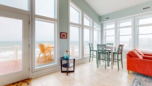 A spacious room with large windows, a dining area, and a red sofa. - Oceanfront Retreat with Premium Views, Roof Top Hot Tub, Private Pool, Game Room and More...! (Surf City)