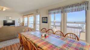 A dining area with a table set for six, a television on a stand, and a view of the beach through large windows. - Oceanfront home with ocean views, beach access, tanning deck & private elevator (Surf City)