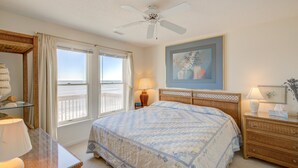 A bedroom with a large bed, a ceiling fan, a window with a view of the beach, and a painting on the wall. - Oceanfront home with ocean views, beach access, tanning deck & private elevator (Surf City)
