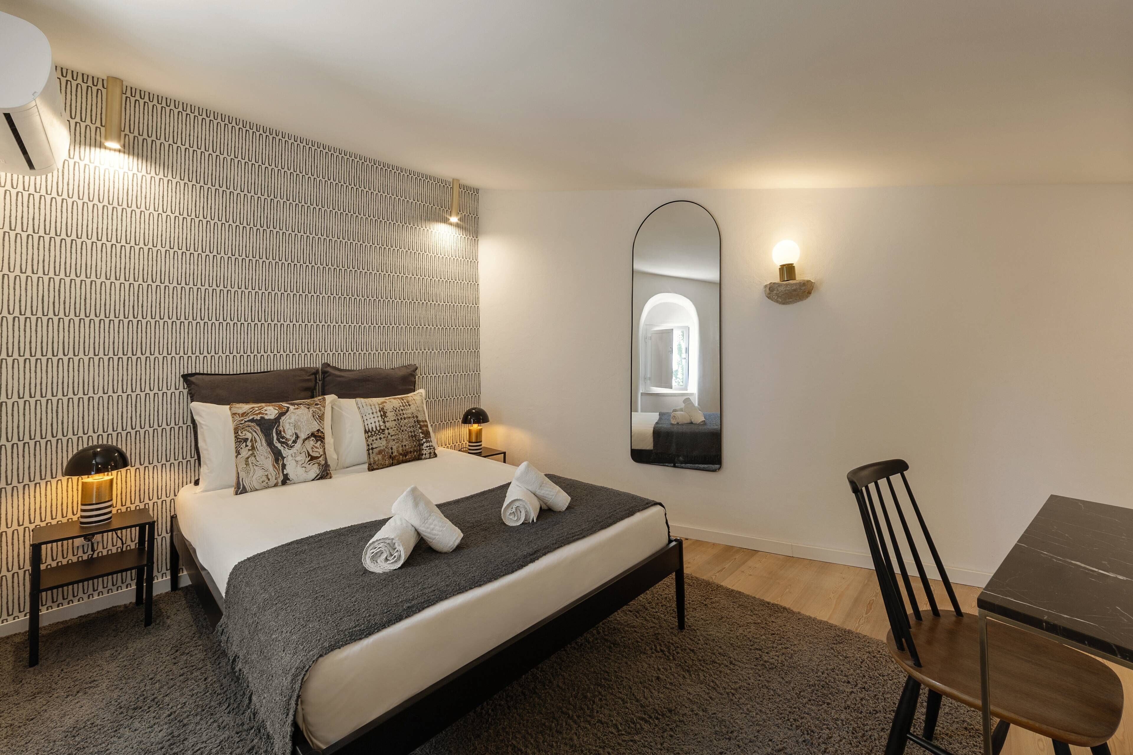 A modern hotel room with a neatly made bed, a bedside table with a lamp, a chair, and a large mirror on the wall.