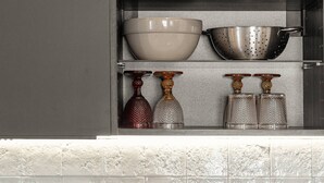 A kitchen with a black countertop, a white tiled backsplash, and a shelf with kitchenware.