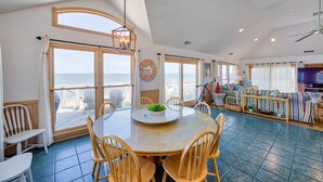 A spacious living area with a large round table, multiple chairs, a television, and a view of the ocean through large windows. - The Sand Jewel: Oceanfront, Private Hot Tub, Pool Access (Avon)