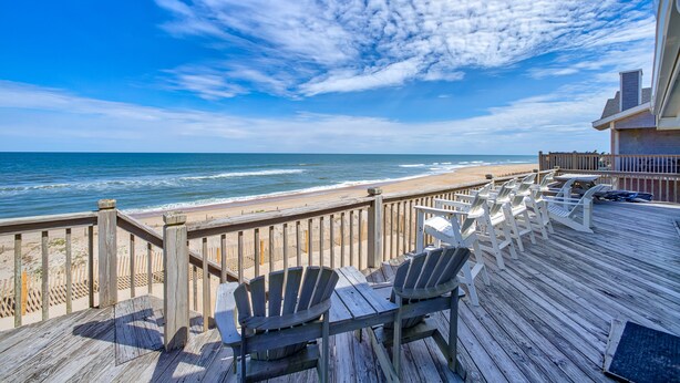 A wooden deck with chairs and tables overlooking a beach with a wooden fence. - The Sand Jewel: Oceanfront, Private Hot Tub, Pool Access (Avon)