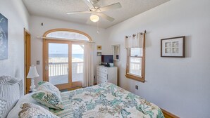 A bedroom with a bed, a ceiling fan, a dresser, a TV, a window with curtains, and a view of the ocean. - The Sand Jewel: Oceanfront, Private Hot Tub, Pool Access (Avon)