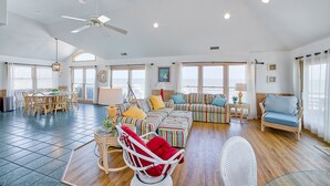 A spacious living room with a dining area, a ceiling fan, and large windows. - The Sand Jewel: Oceanfront, Private Hot Tub, Pool Access (Avon)